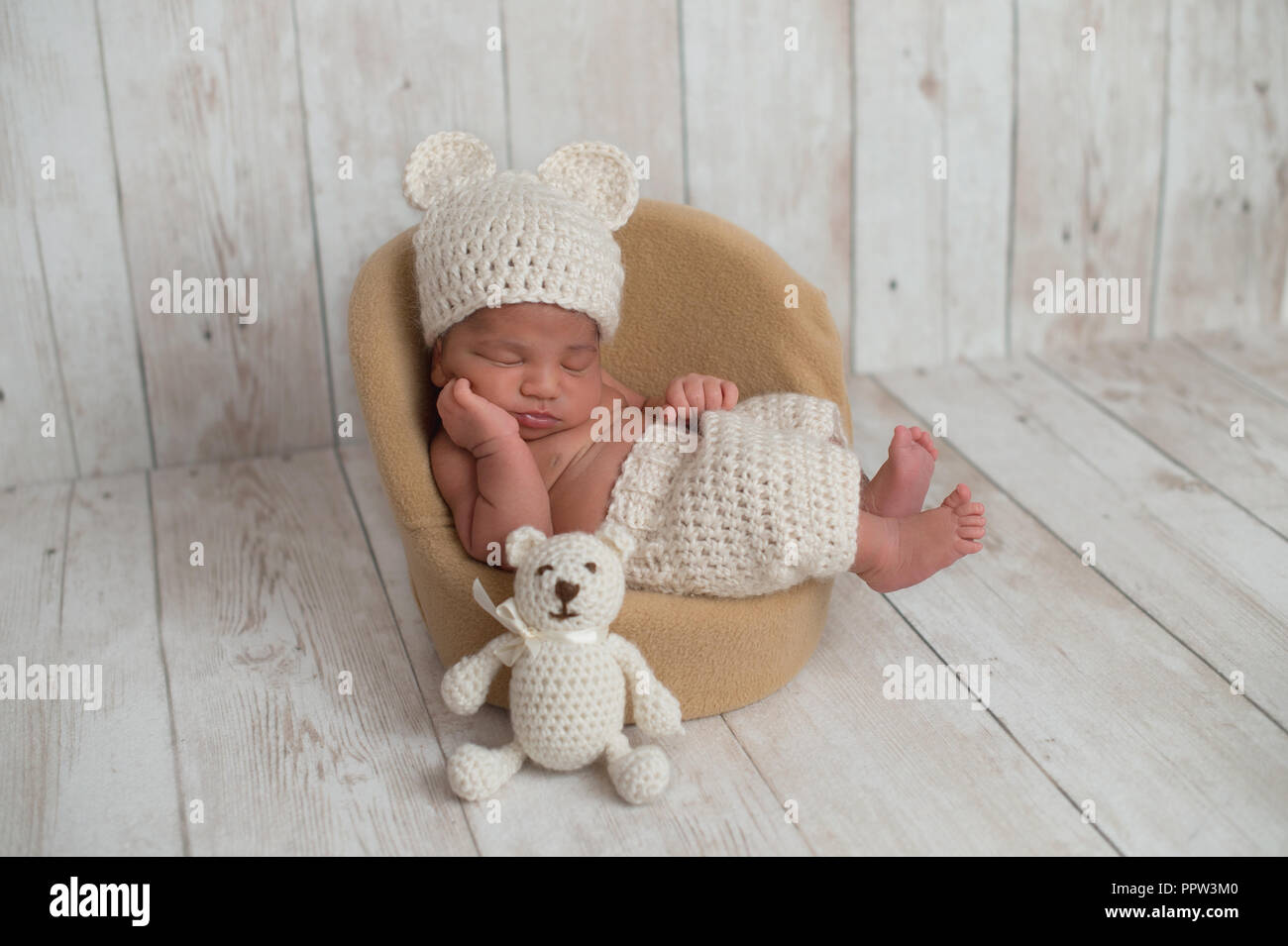 Nine day old newborn baby boy wearing cream colored, crocheted, bear hat and shorts. He is sleeping on a tiny chair with a matching stuffed bear toy n Stock Photo