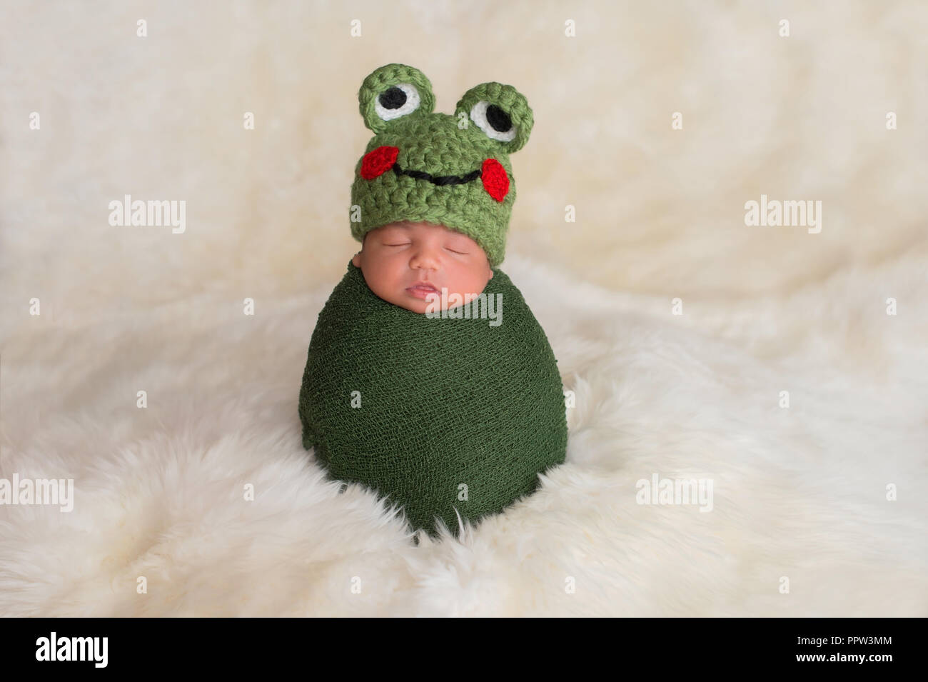 Nine day old newborn baby boy wearing a green frog hat. He is sleeping upright while swaddled in a stretch wrap. Stock Photo