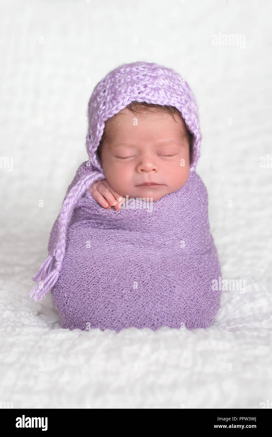 A two week old newborn baby girl wearing a lavender, crocheted bonnet. She is sleeping upright while swaddled in a light purple stretch wrap. Stock Photo