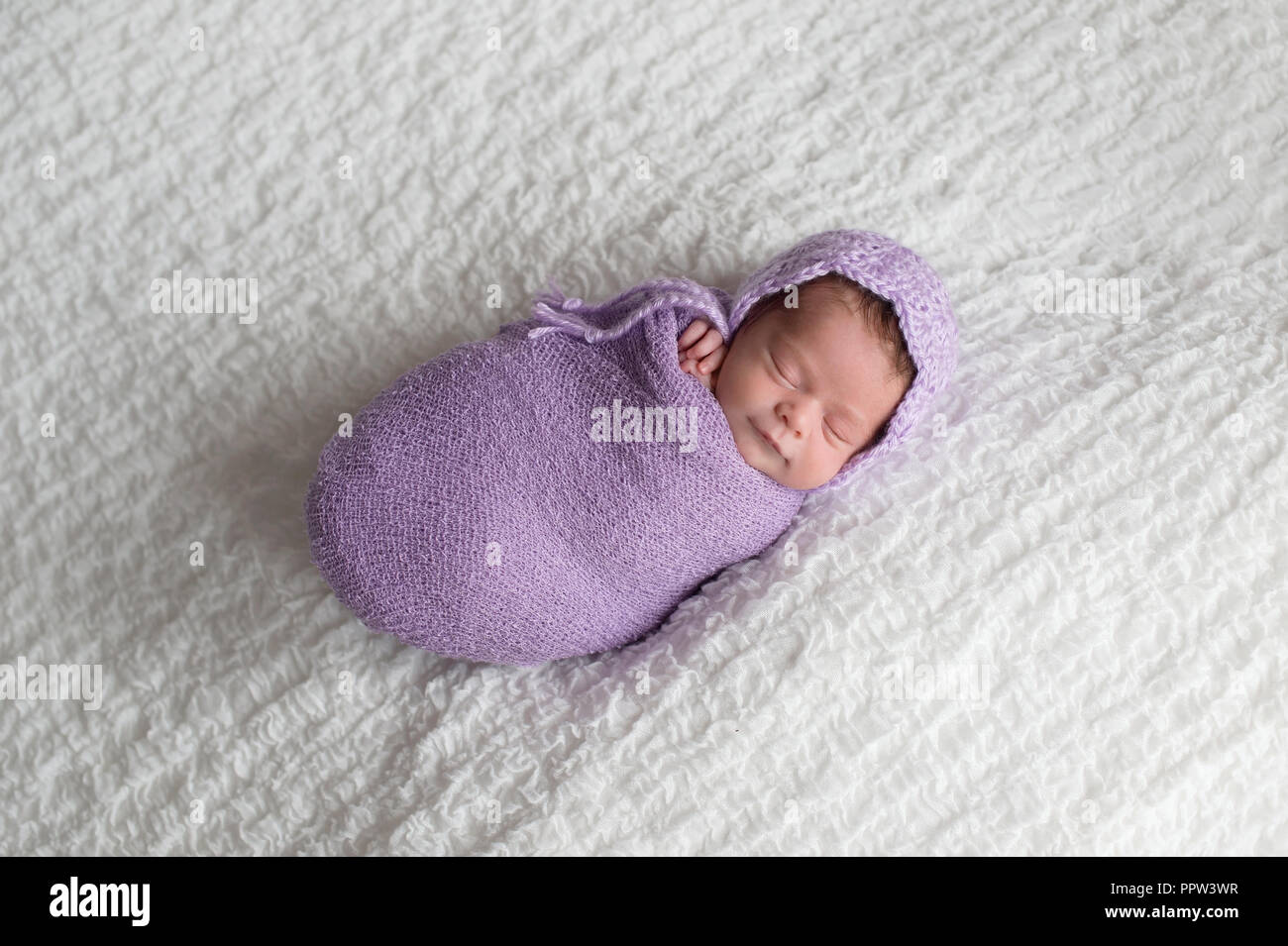A smiling, two week old newborn baby girl swaddled in a lavender wrap and wearing a crocheted bonnet. Shot in the studio on a white blanket. Stock Photo