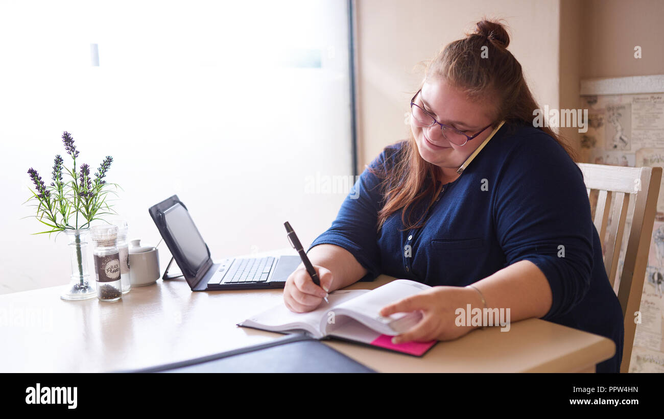 Plus size business woman busy multitasking at her local coffee shop to get as much work done as possible early in the morning, as she writes in her jo Stock Photo