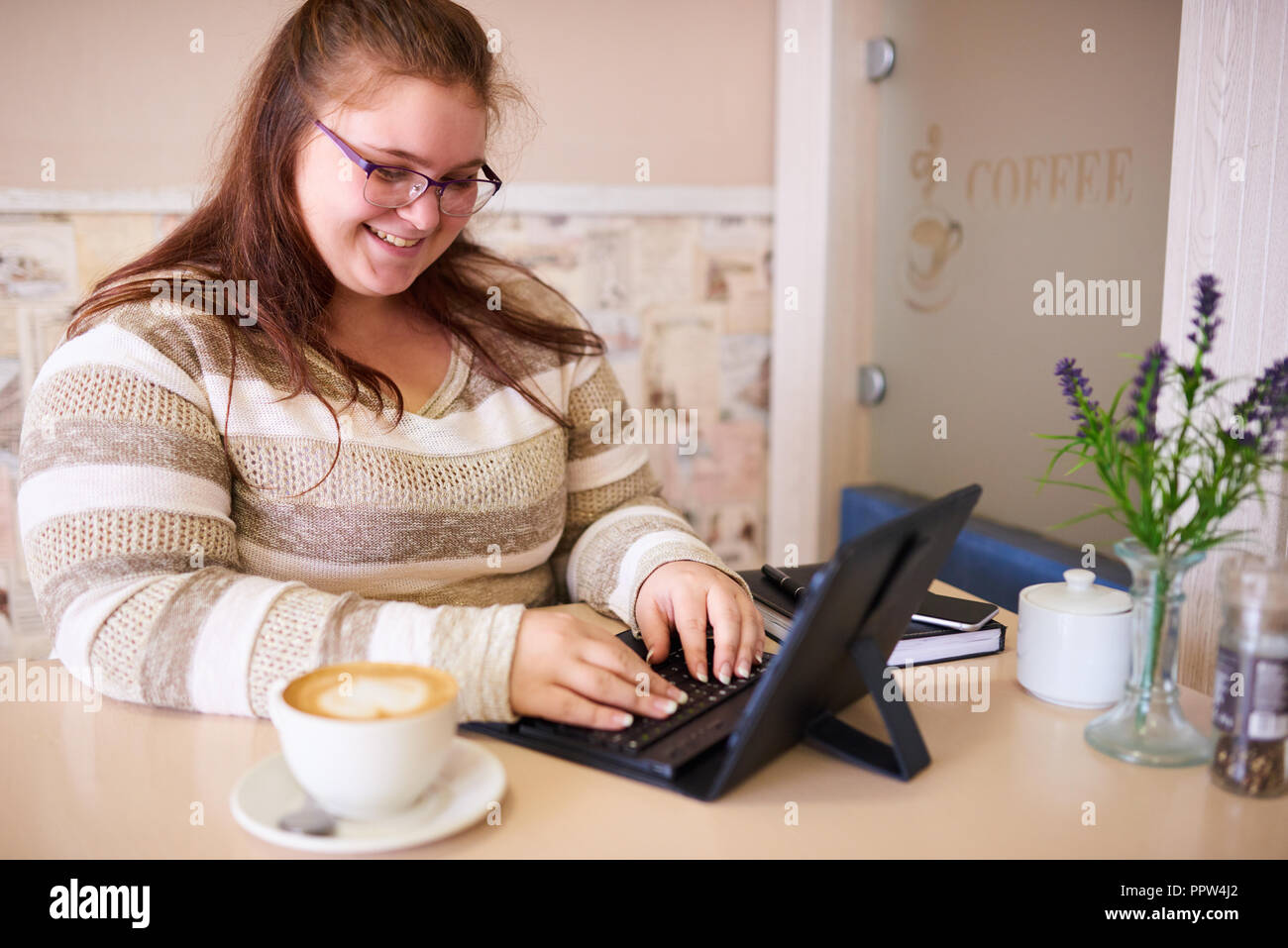 Plus size caucsian woman happily smiling while working on her tablet and keyboard in a bright cafe with a morning cup of coffee to be more productive. Stock Photo