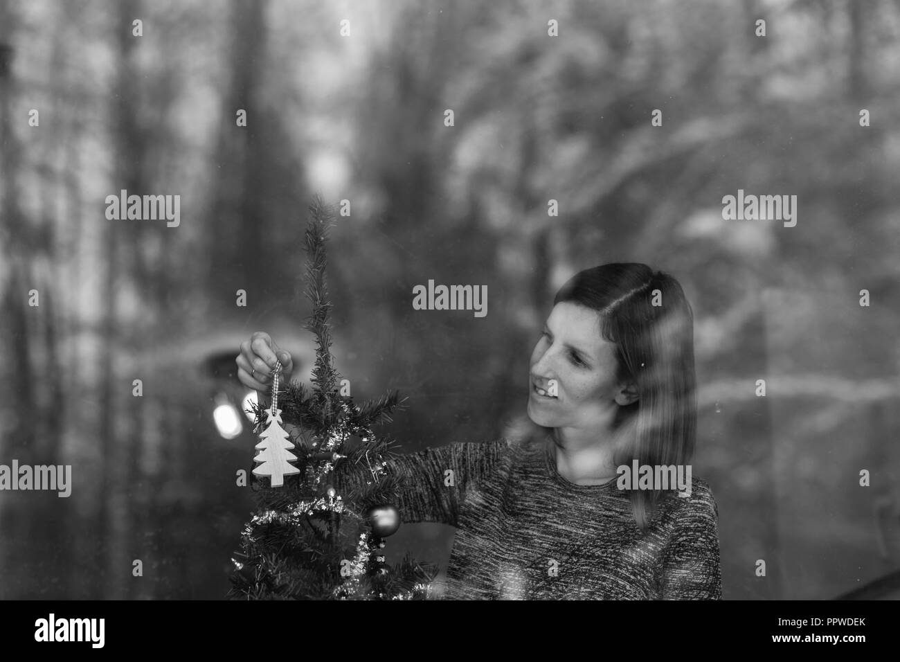 Monochrome image of a young woman placing wooden holiday ornament on a Christmas tree. View through a window. Stock Photo