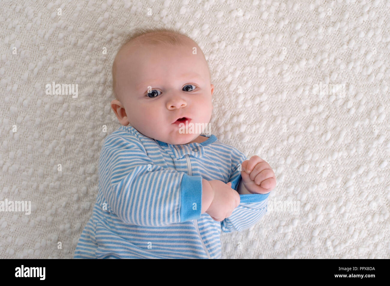 A 2 month old baby boy lying on his back on a white blanket. He is wearing blue and white striped pajamas and is looking at the camera. Stock Photo