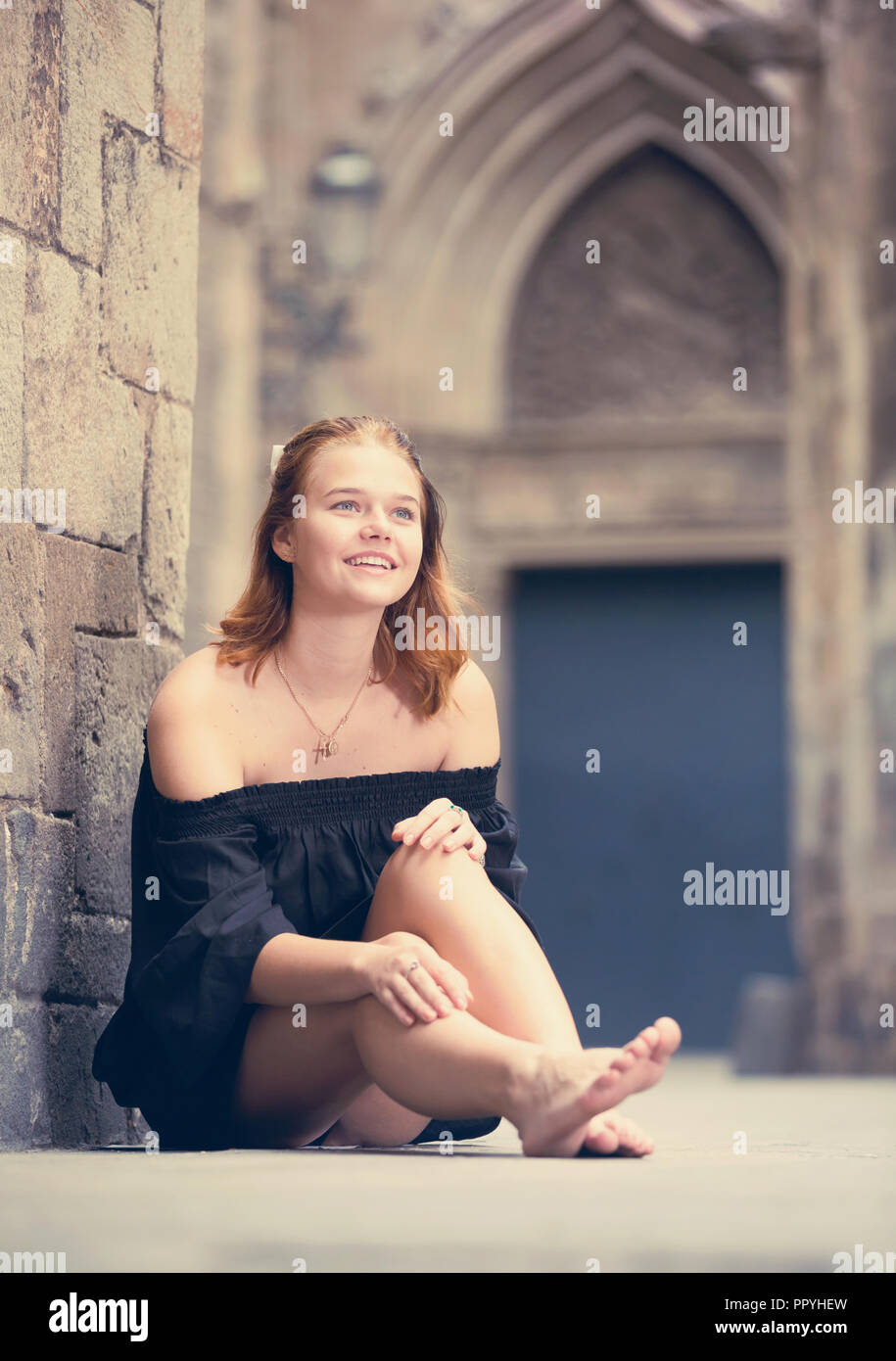Positive Young Girl Sitting Barefoot Near The Stone Wall At The Street positive-young-girl-sitting-barefoot-near-the-stone-wall-at-the-street