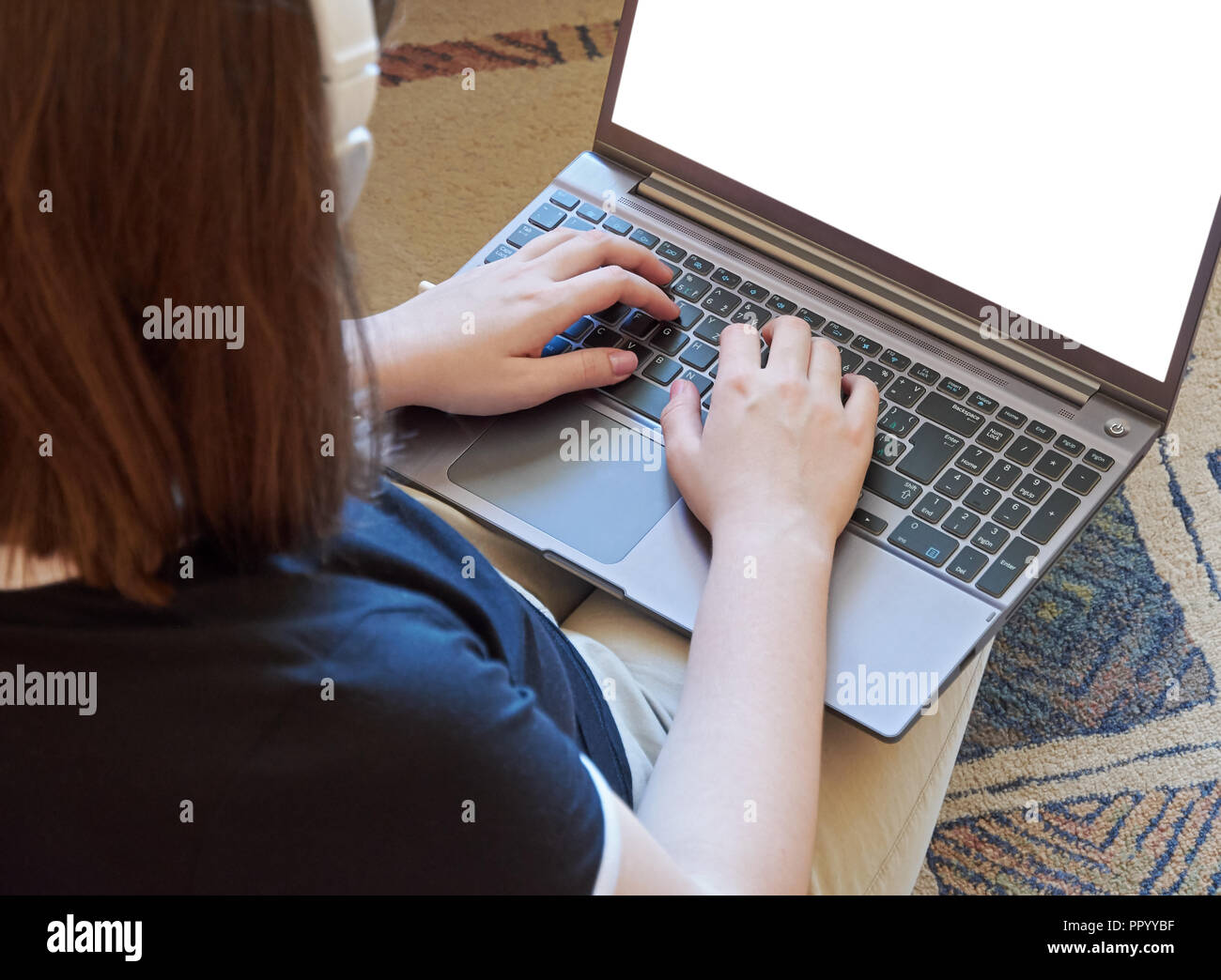 Young woman with laptop on her lap, writes on keyboard and looking at white display, with headphones. Sits on the floor on the carpet Stock Photo