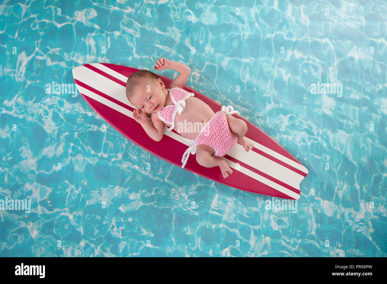 Two week old newborn baby girl lying on a tiny, pink and white surfboard. She is wearing a pink, crocheted bikini. Stock Photo