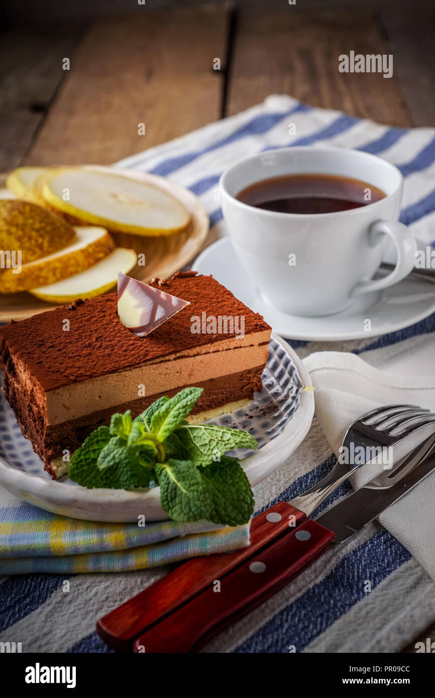 A slice of Chocolate cake on a plate, a cup of tea, lemon, sprig of mint, and slices of pear on background, vertical. Stock Photo