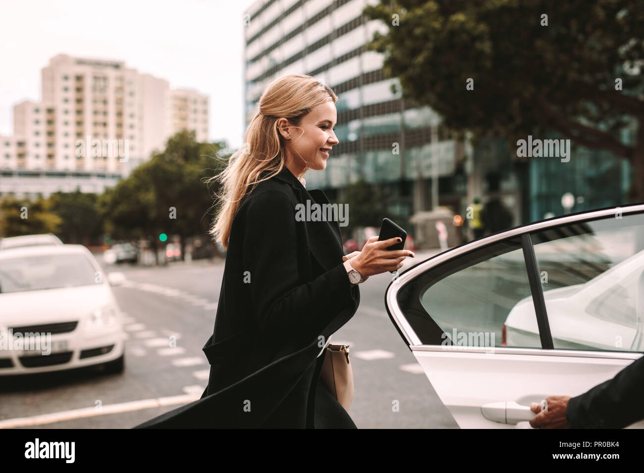 Smiling businesswoman getting into a taxi. Female commuter entering a taxi with driver opening door. Stock Photo