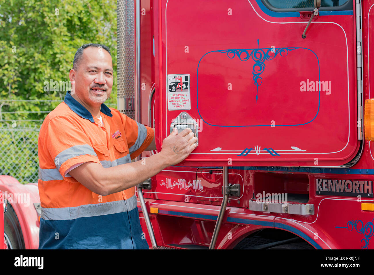 A happy Samoan truck driver in hi-vis clothing, climbing into a the cab of a red semi trailer prime mover truck Stock Photo