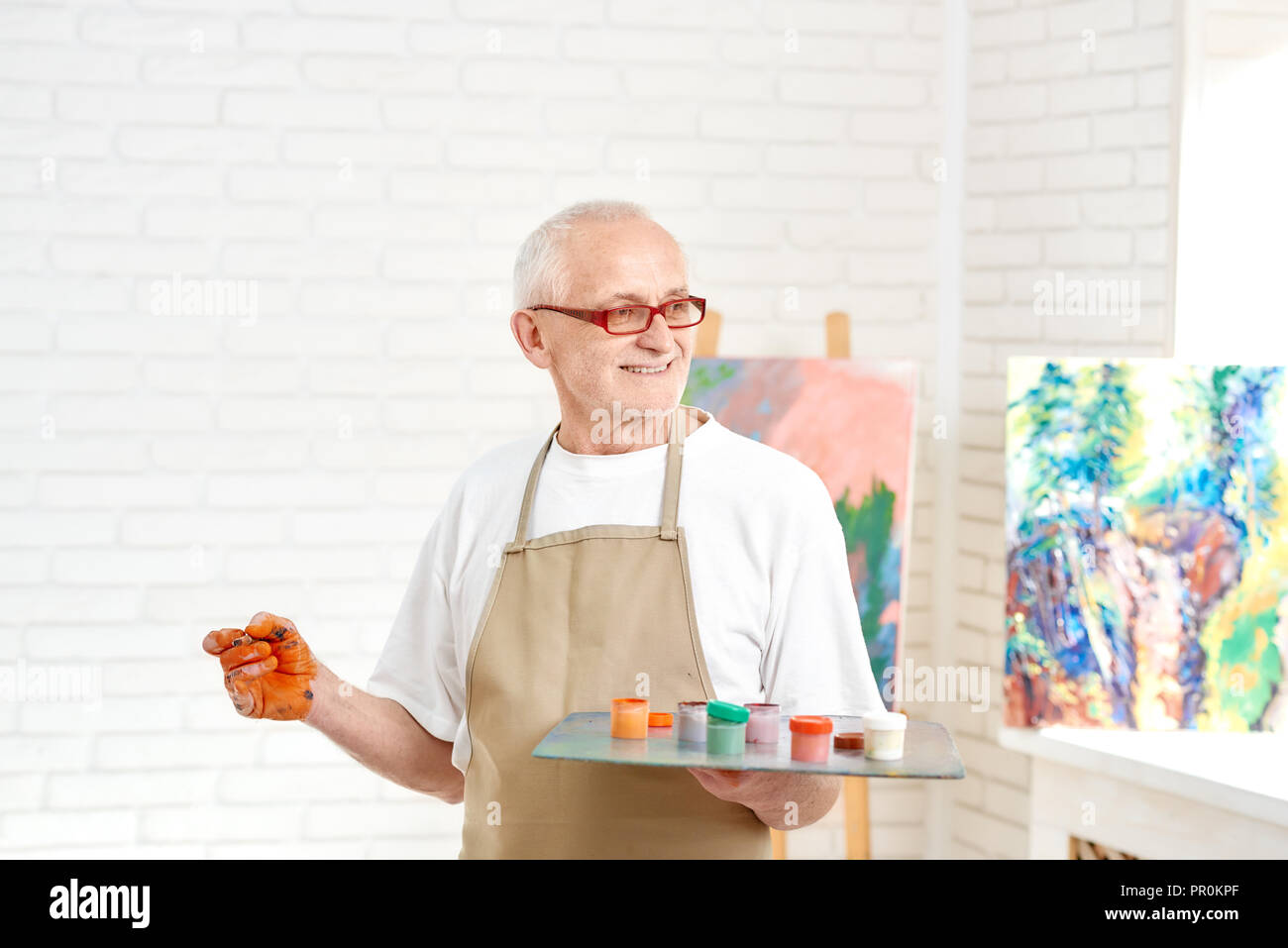 Senior male painter, standing with palette of colors in hand, picking up paint to picture, other hand contaminated with orange paint. Handsome man wearing in white t-shirt and apron, looking at work. Stock Photo