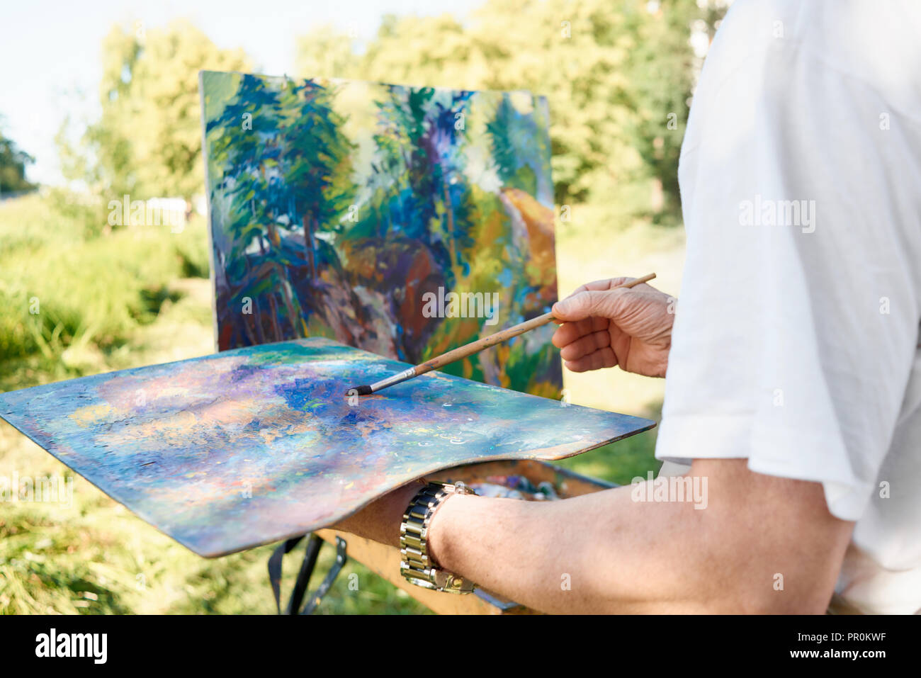 Close-up of artist hand holding painting brush and palette of colors against almost finished picture at park. Easel and canvas with picture standing against lake on open air. Concept of hobby. Stock Photo