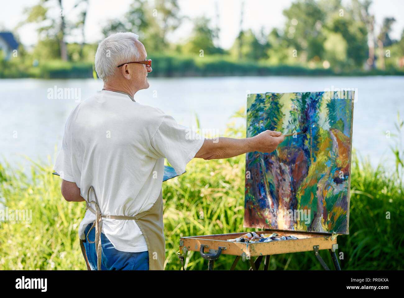 Rear view of creative painter paints colorful abstract painting on open air. Senior painter, wearing in apron and modern glasses, using palette with paints working on his new masterpiece against lake. Stock Photo