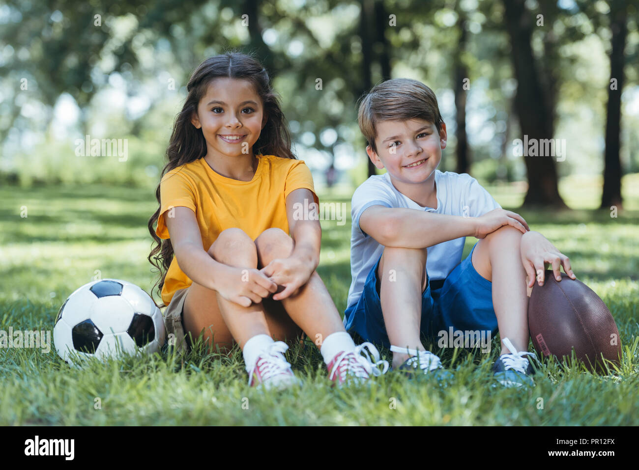 cute happy children with soccer and rugby balls sitting and smiling at camera in park Stock Photo