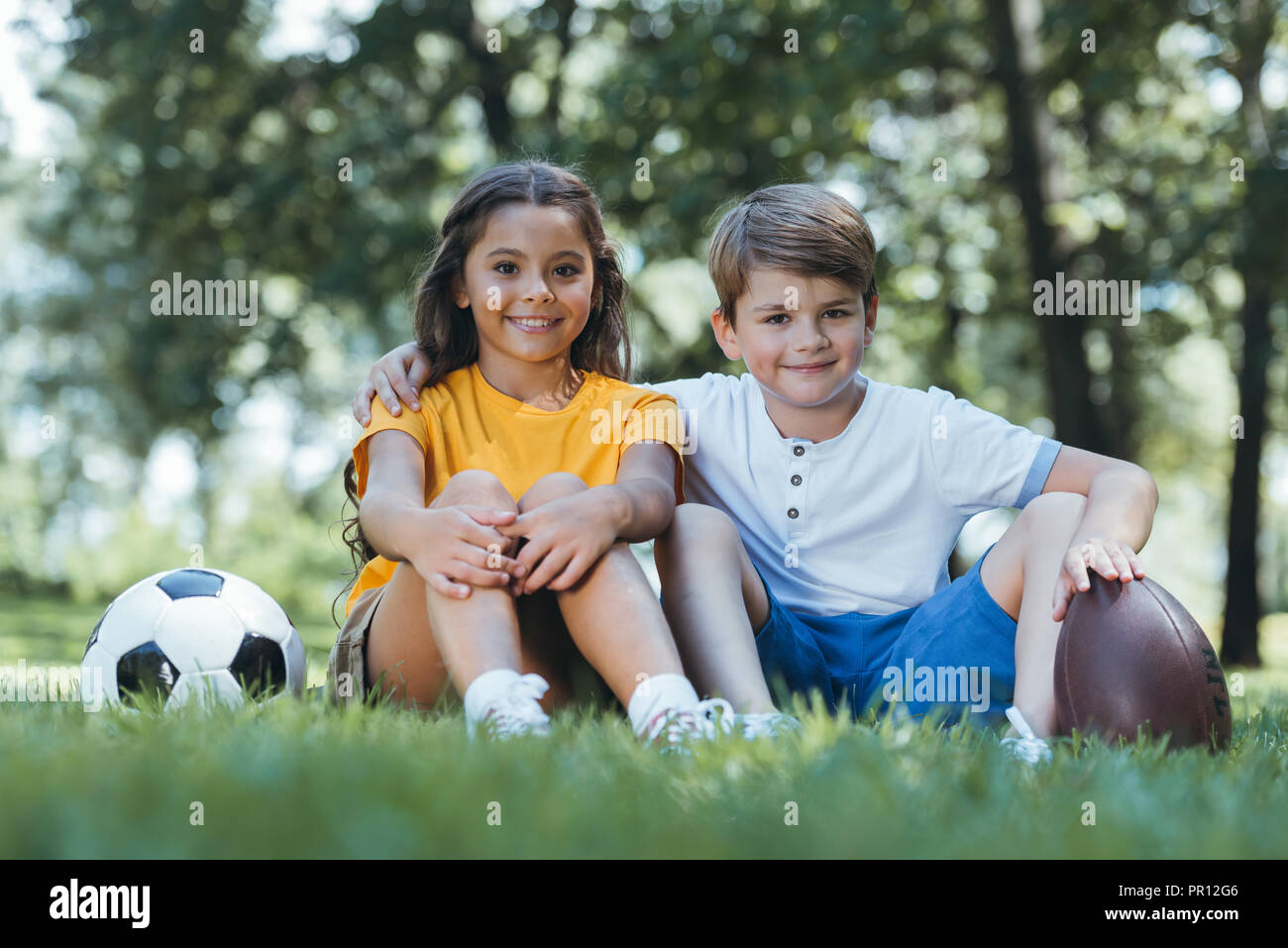 cute happy kids with soccer and rugby balls sitting on grass and smiling at camera Stock Photo