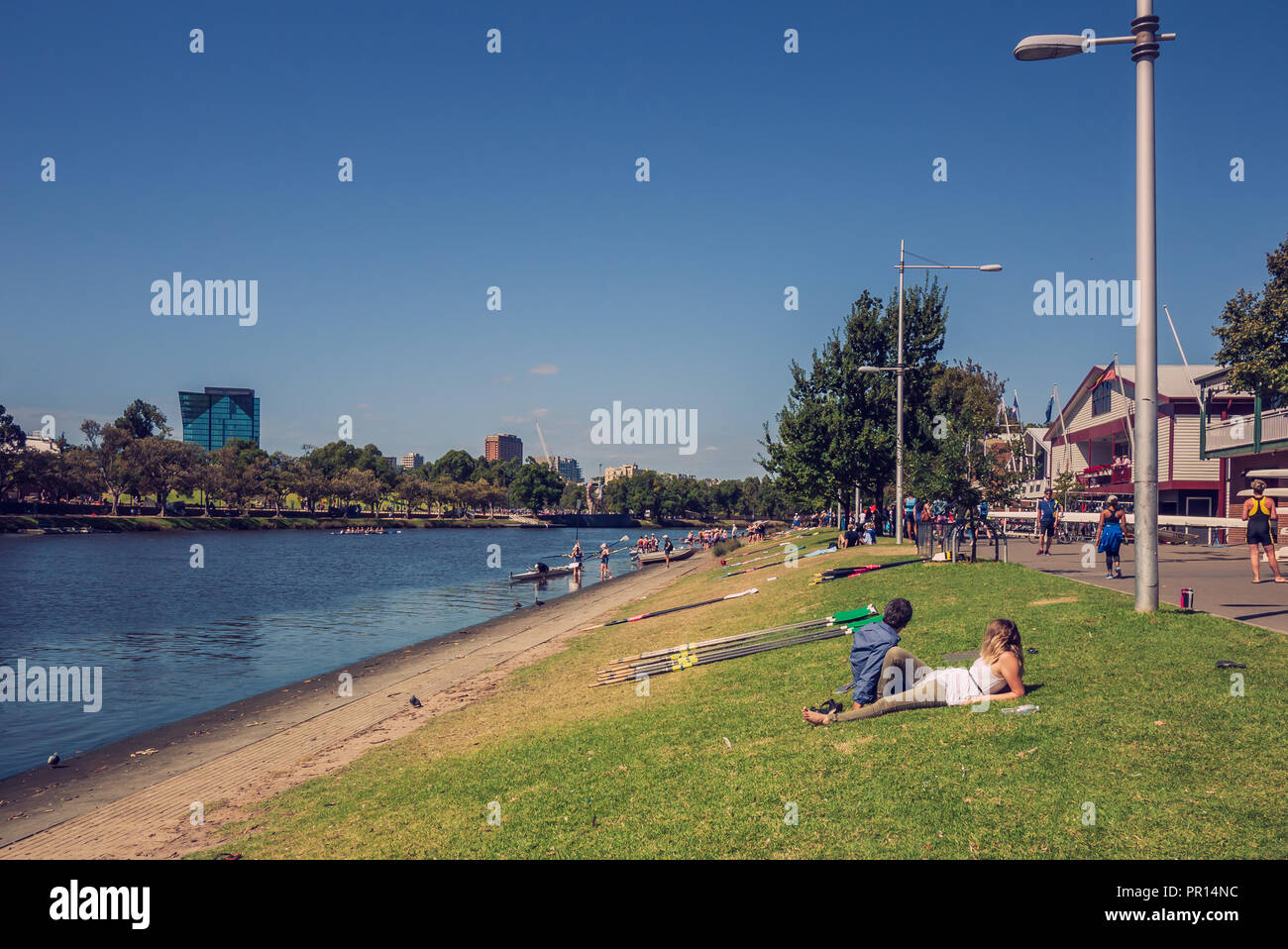 Man and woman laying on the lawn at the river bank of Yarra. There are lots of rowing clubhouses and the players are preparing the boat on the water. Stock Photo