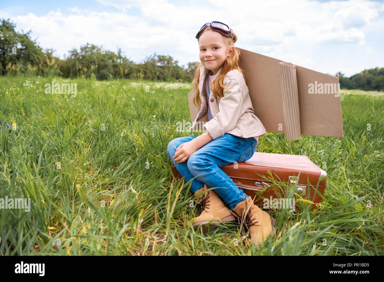 smiling cute kid in pilot costume sitting on retro suitcase in summer ...