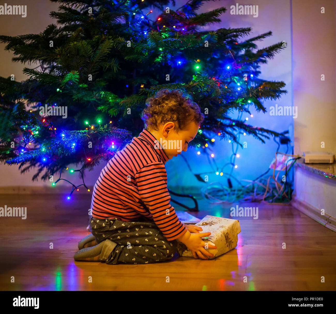 Cute toddler opens gifts on the floor near the Christmas tree (evening mystery lighing) Stock Photo