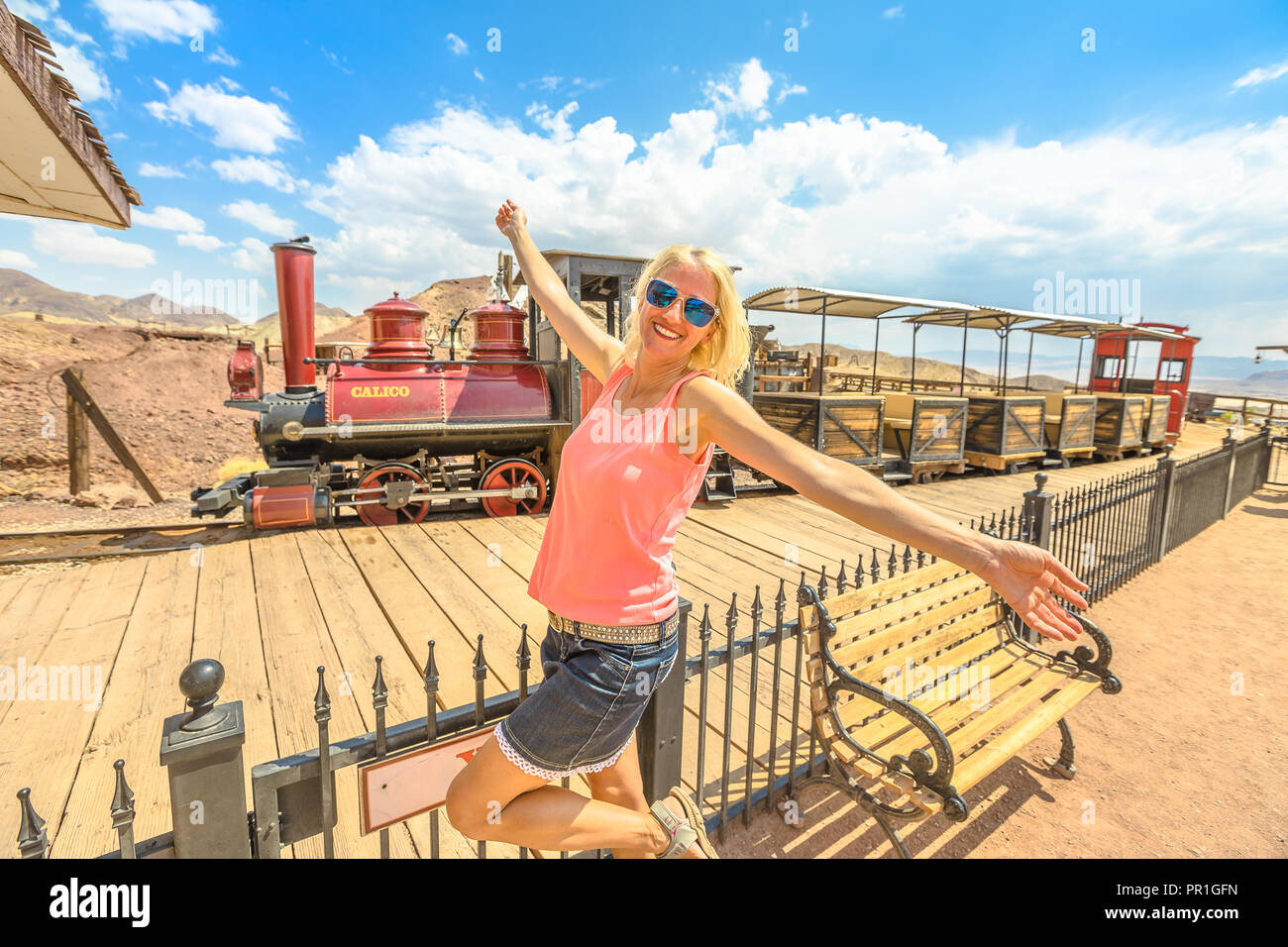 Carefree woman with open arms at Calico Station with old steam train ready for a tour through old mines of Calico Ghost Town, San Bernardino County, California, United States. Stock Photo