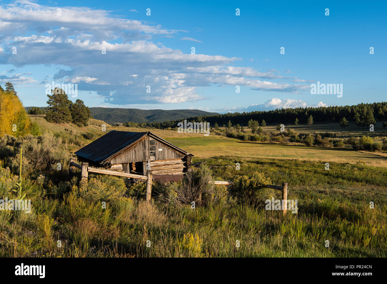 Old rustic log cabin overlooking a landscape of ranchland, fields, forest, and hills in northern New Mexico in late afternoon sunlight Stock Photo