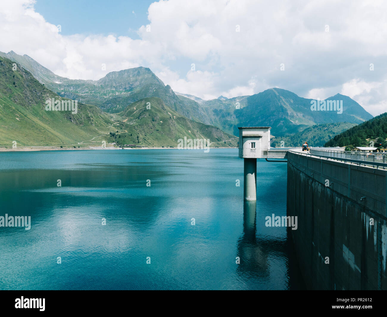 Dam in the Alps blocking the water of the lake Ritom (Ritòm) in Switzerland. Mountains in the background, different-shaded water and cloudy sky. Stock Photo