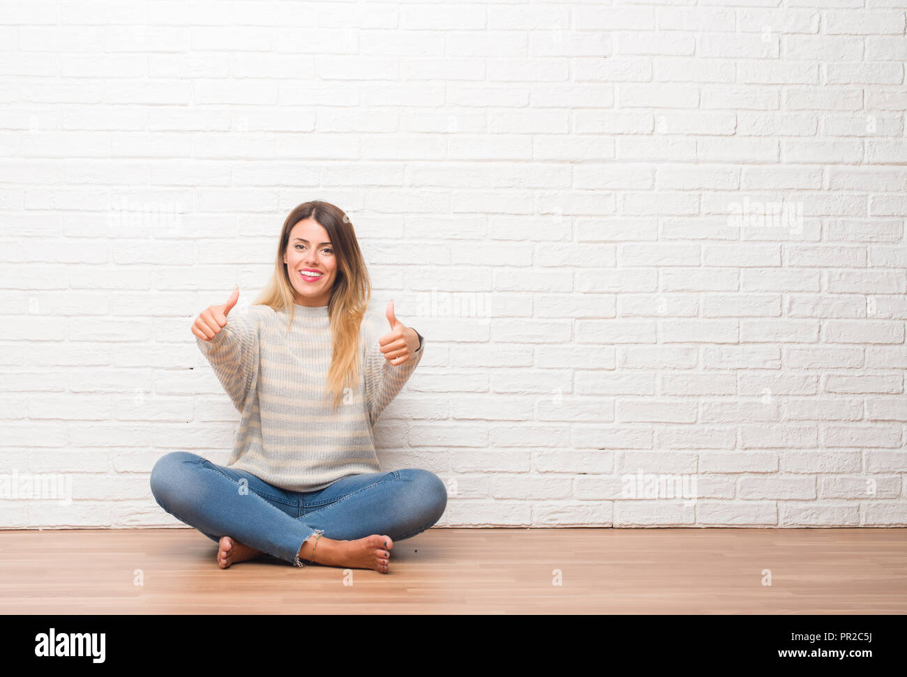 Young adult woman sitting on the floor over white brick wall at home ...
