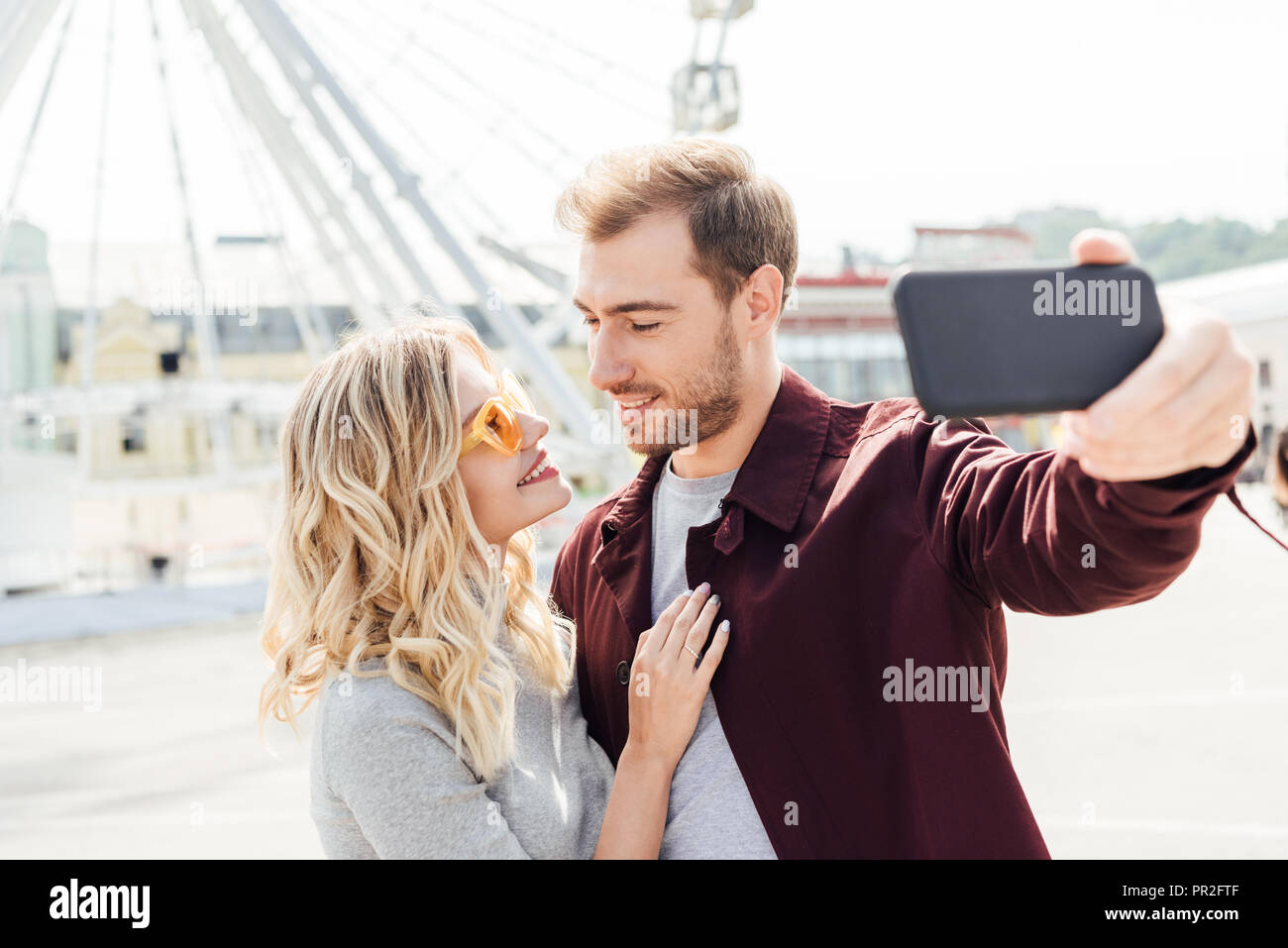 smiling couple in autumn outfit looking at each other and taking selfie with smartphone in city Stock Photo