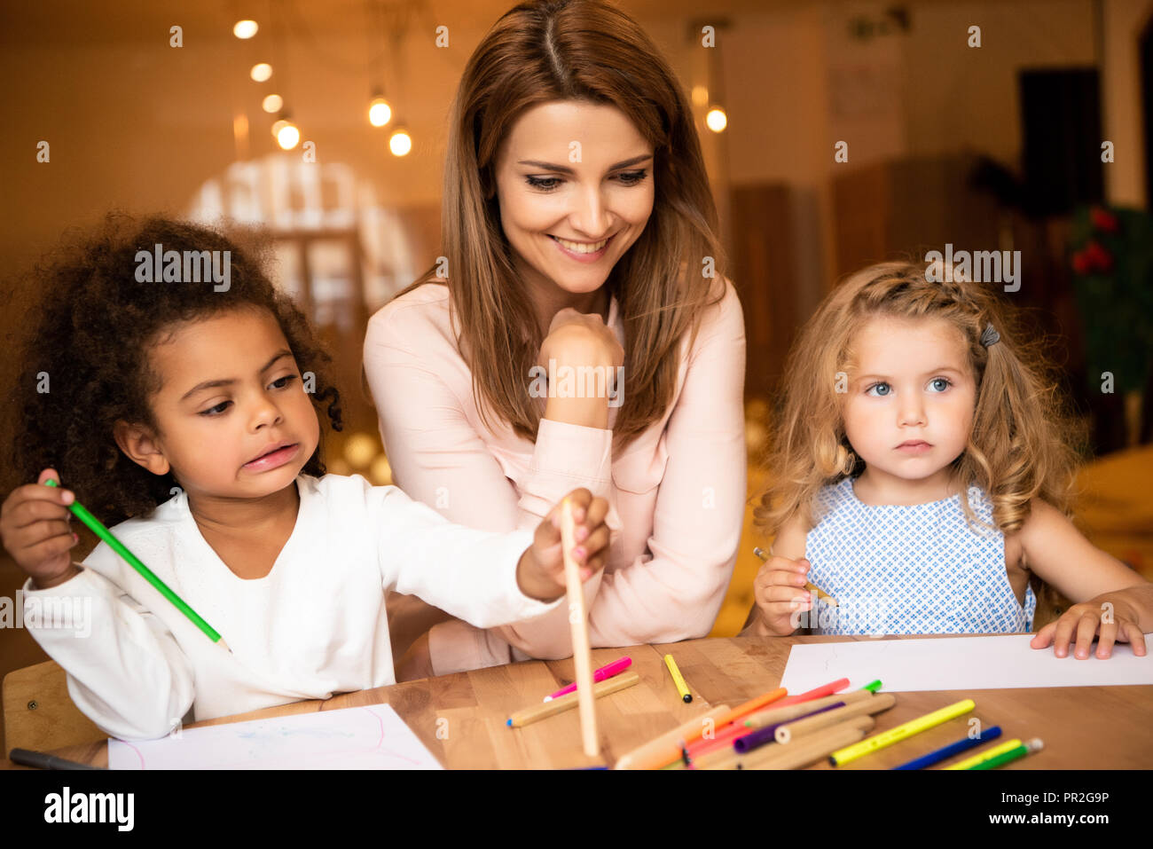 cheerful educator sitting near multicultural kids drawing in ...
