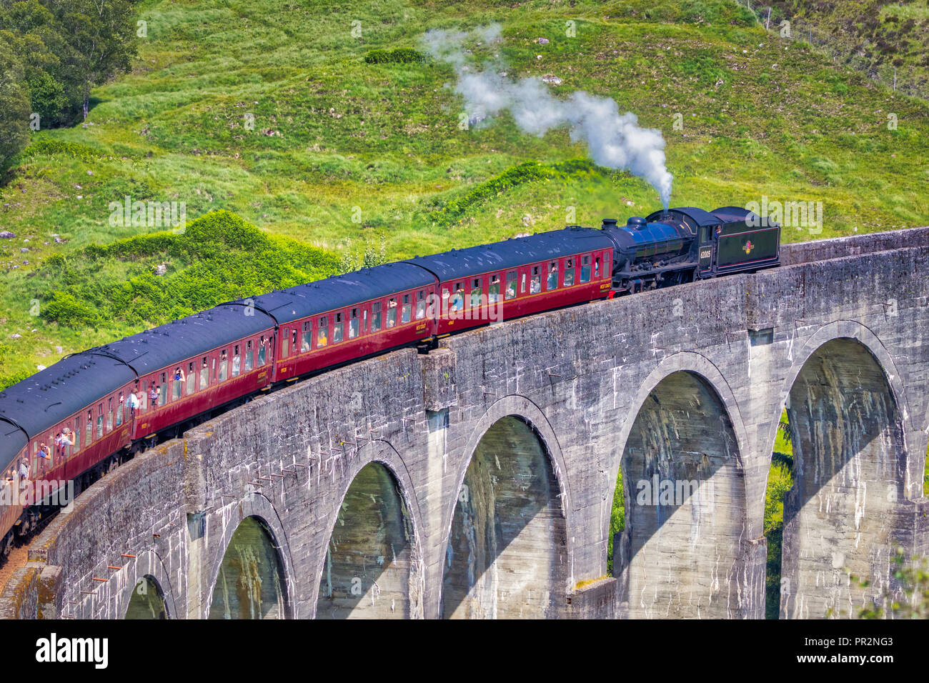 Close shot of Jacobite train travelling over the Glenfinnan Viaduct with a green hillside behind it and steam coming out of the locomotive Stock Photo