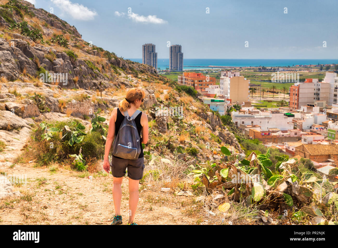 A woman hiking with a backpack up a rocky mountain with cacti and Cullera, Spain and the sea can be seen in the distance Stock Photo