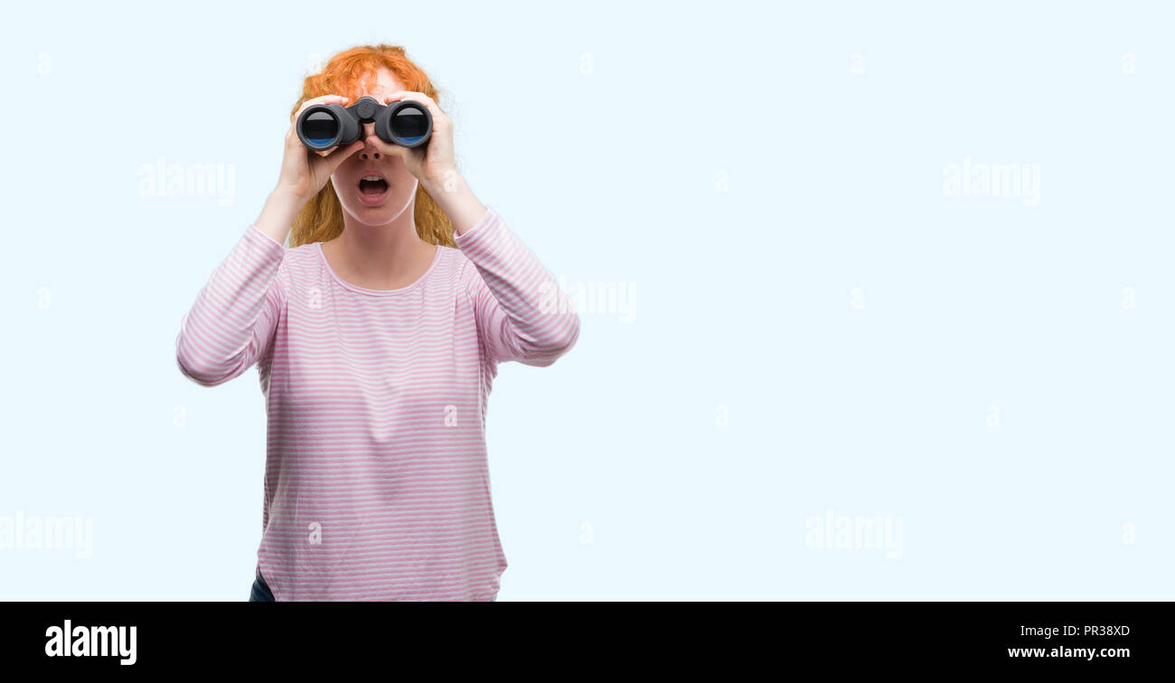 Young redhead woman looking through binoculars scared in shock with a ...