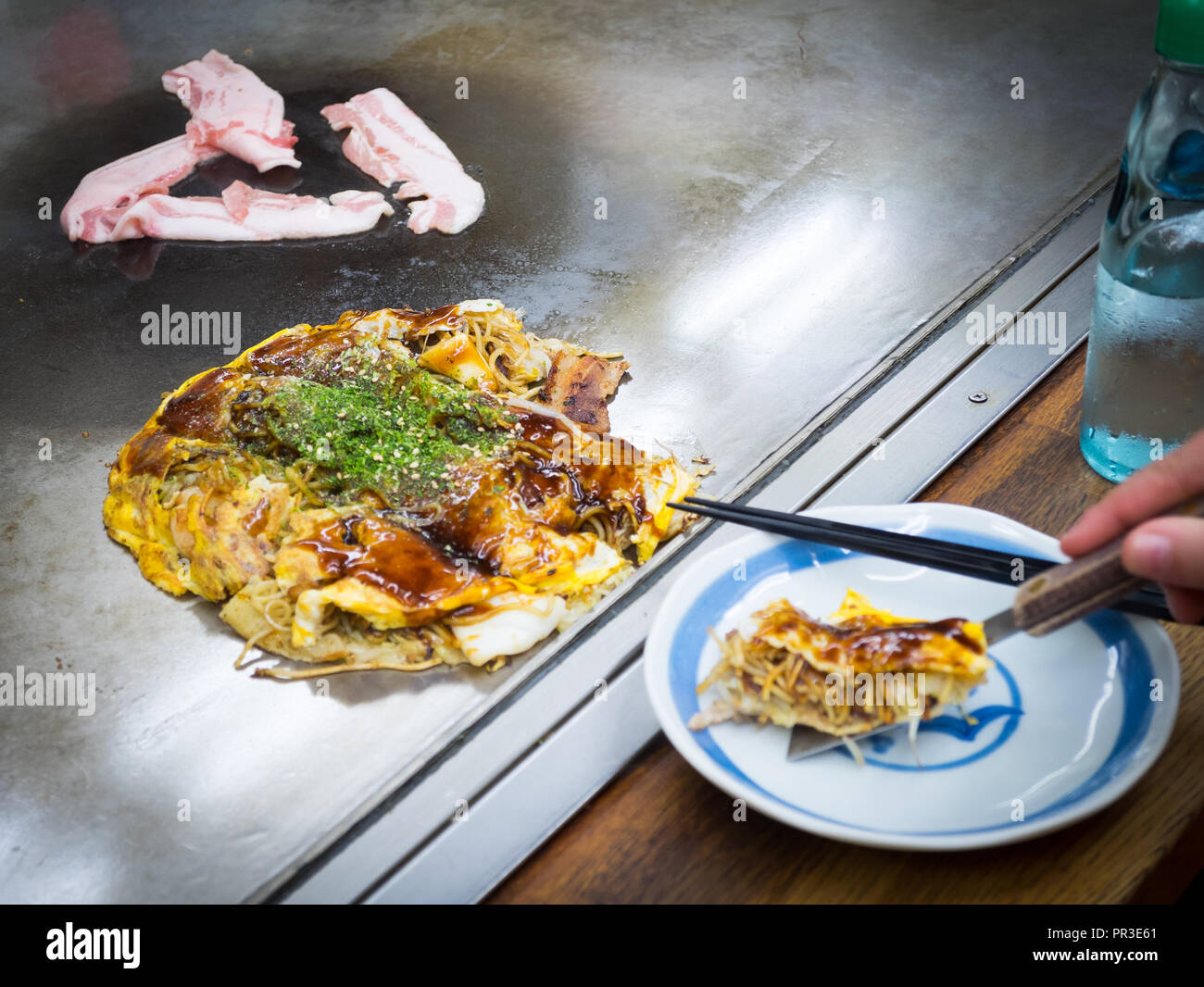Hiroshima-style okonomiyaki with cabbage, udon noodles, squid, bacon, and beef on a hot grill at Okonomimura in Hiroshima, Japan. Stock Photo