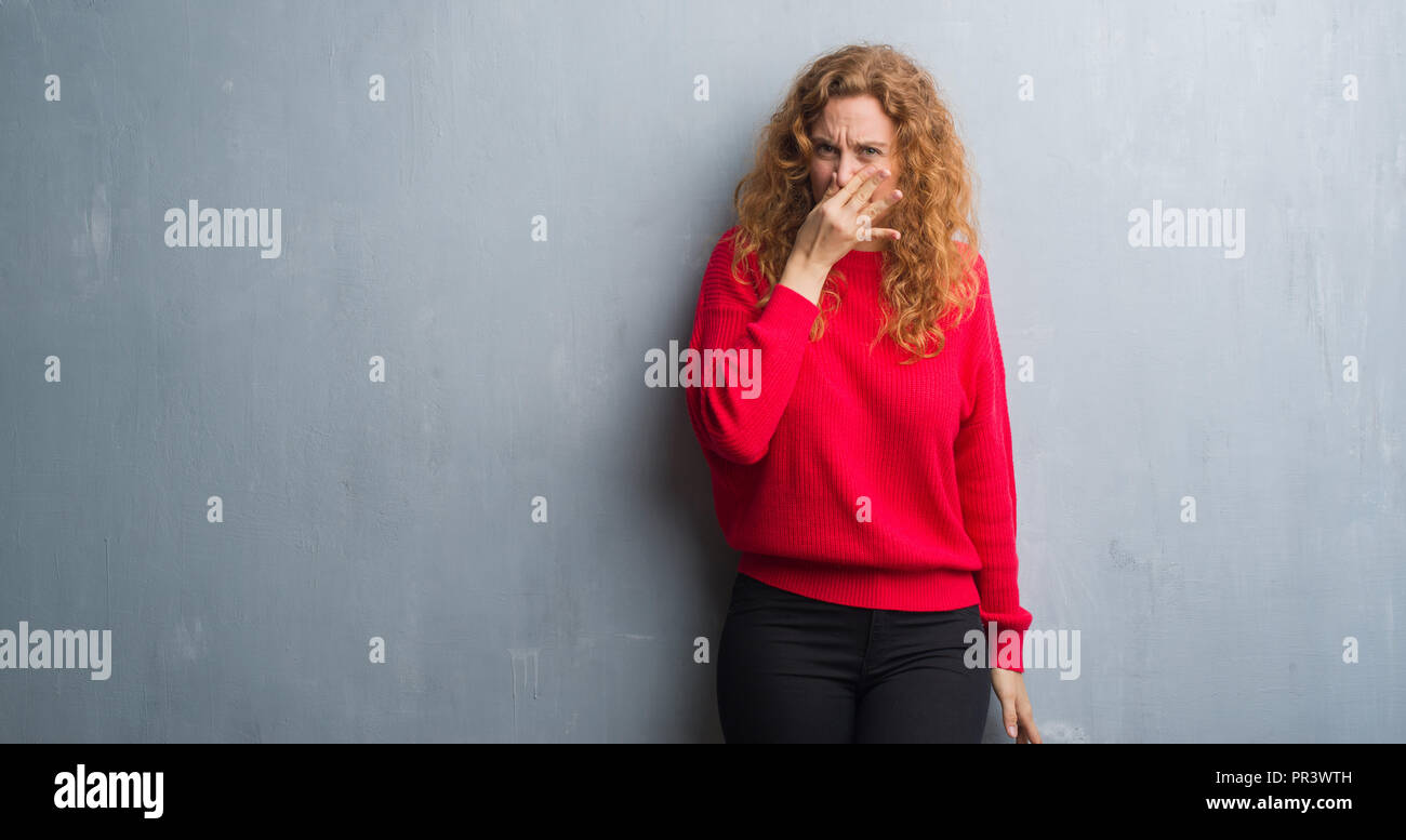Young redhead woman over grey grunge wall wearing red sweater smelling ...