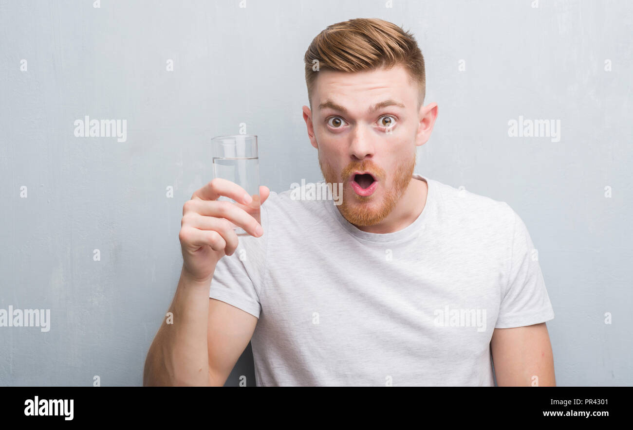 Young redhead man over grey grunge wall drinking water scared in shock ...
