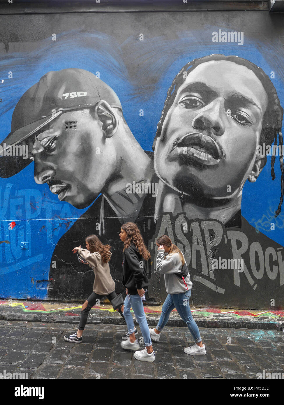 Vertical shot of three teenage girls walking in front of a beautiful portrait of two young black men painted on wall in Hosier Lane in Melbourne. Stock Photo