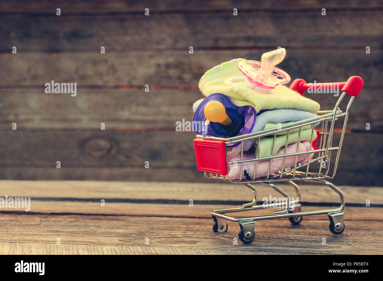 Shopping cart with clothing and baby pacifiers on the old wood background. Toned image. Stock Photo