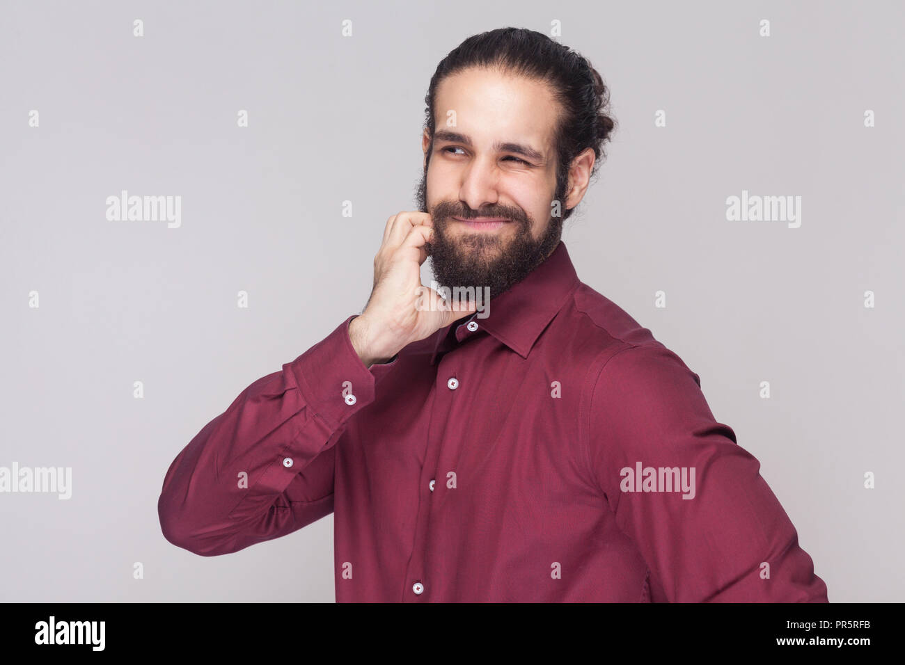 Portrait of confused handsome man with dark collected long hair and beard in red shirt standing and touching his face, thinking and looking away. indo Stock Photo