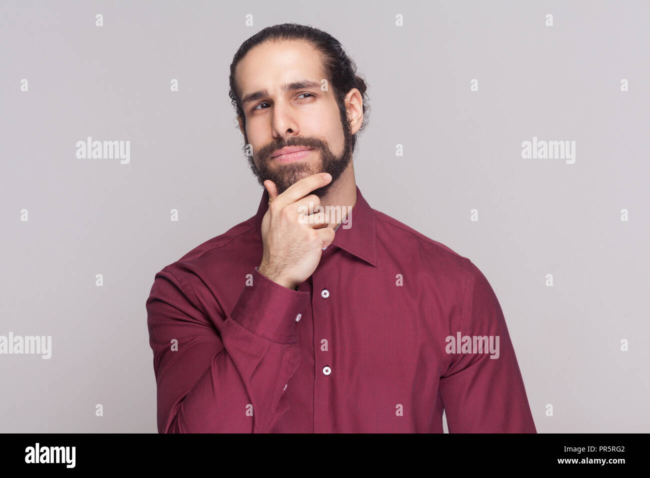 Portrait of thoughtful handsome man with dark collected long hair and beard in red shirt standing and touching his chin, thinking and looking away. in Stock Photo