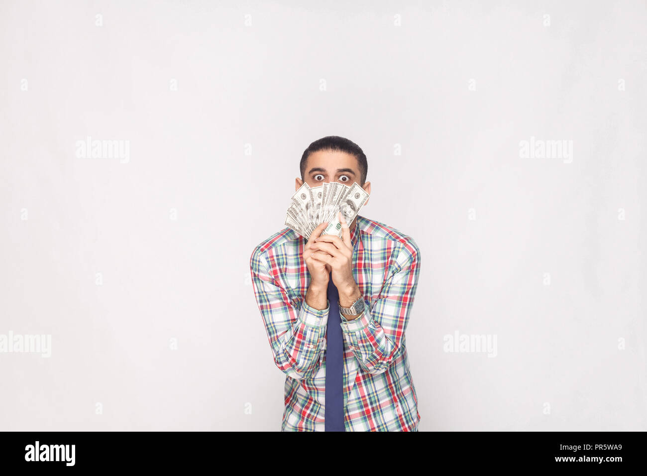 Portrait of rich handsome young adult businessman in colorful checkered shirt with blue tie standing and showing fan of dollars with big crazy eyes. I Stock Photo