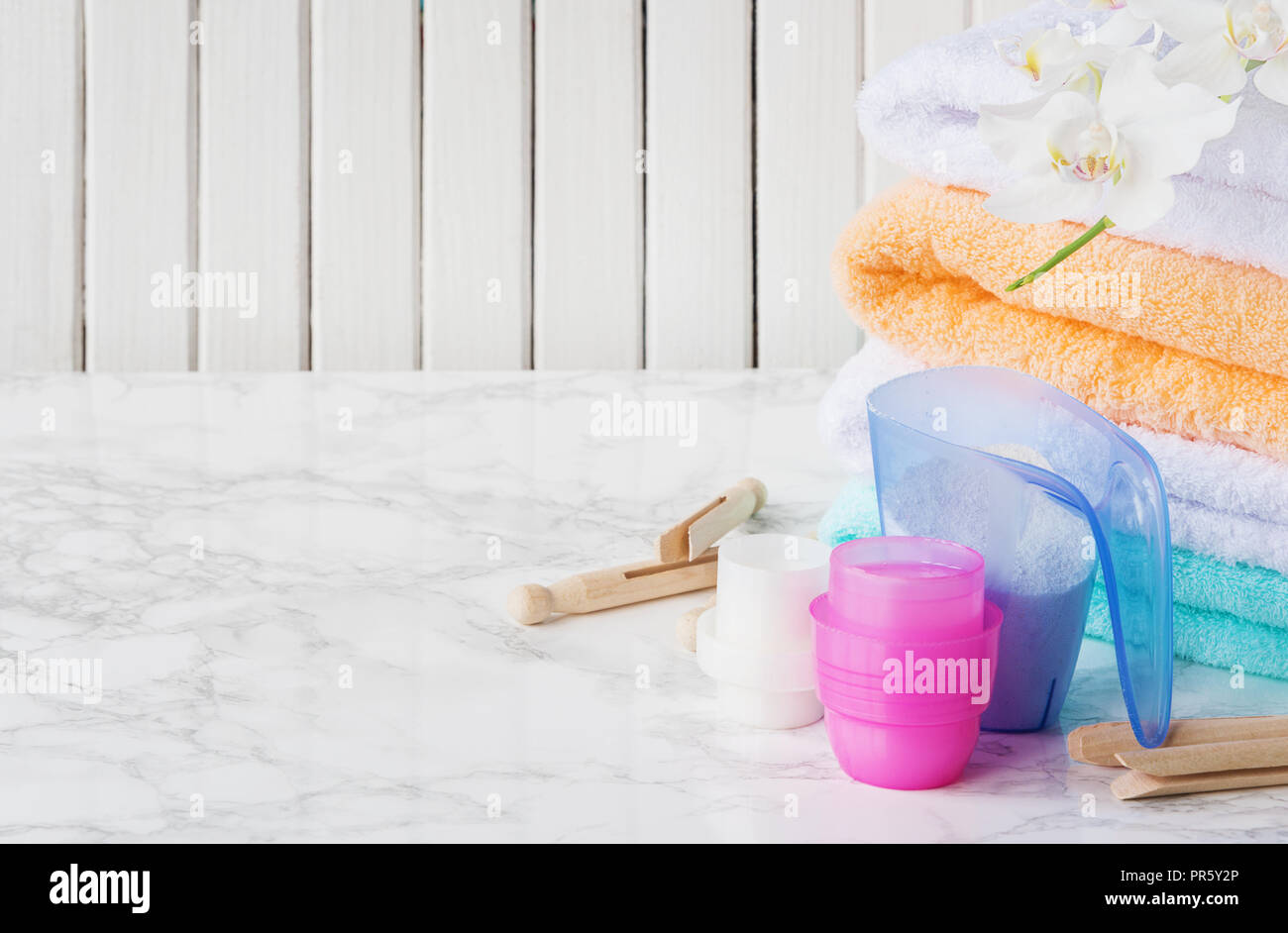 Blue plastic measuring vessel with washing powder, containers with detergent, stack of white terry towels, wooden clothespins and white orchid flower  Stock Photo