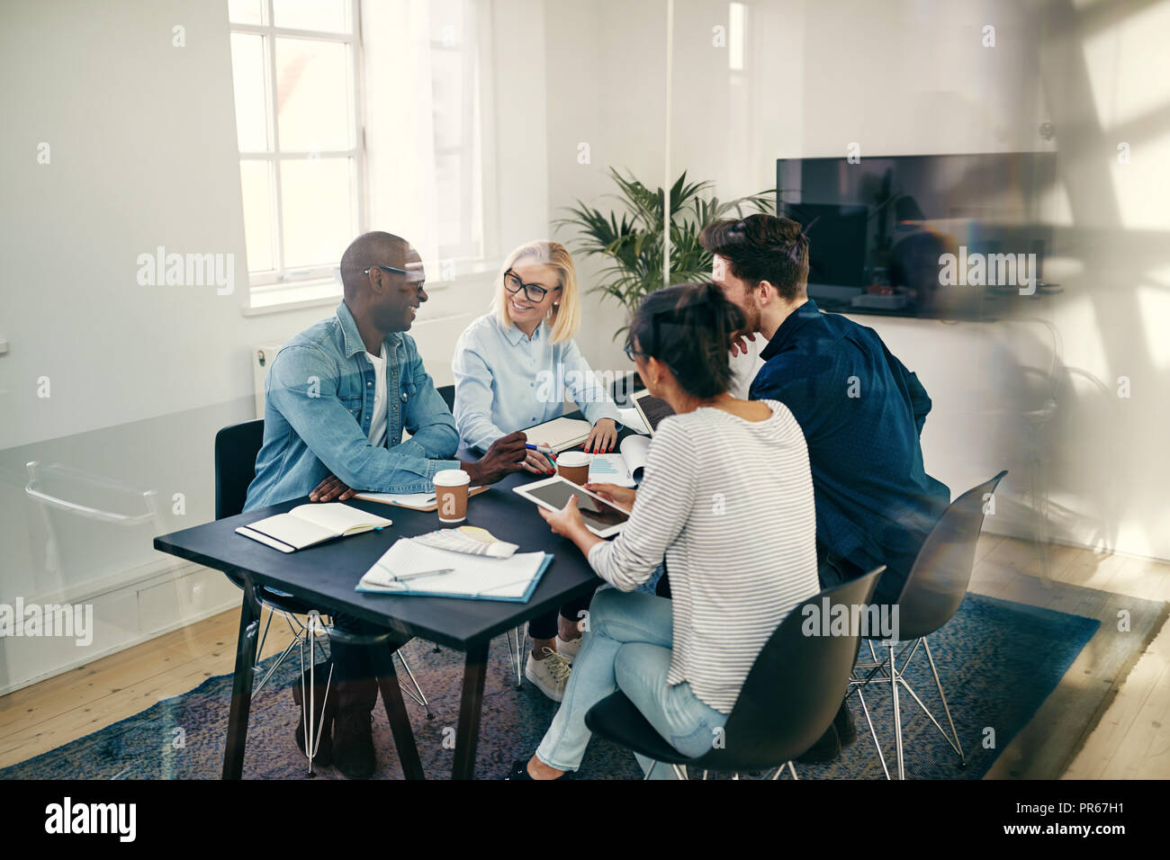 Diverse group of smiling young businesspeople talking together during a meeting inside of a glass walled office boardroom Stock Photo