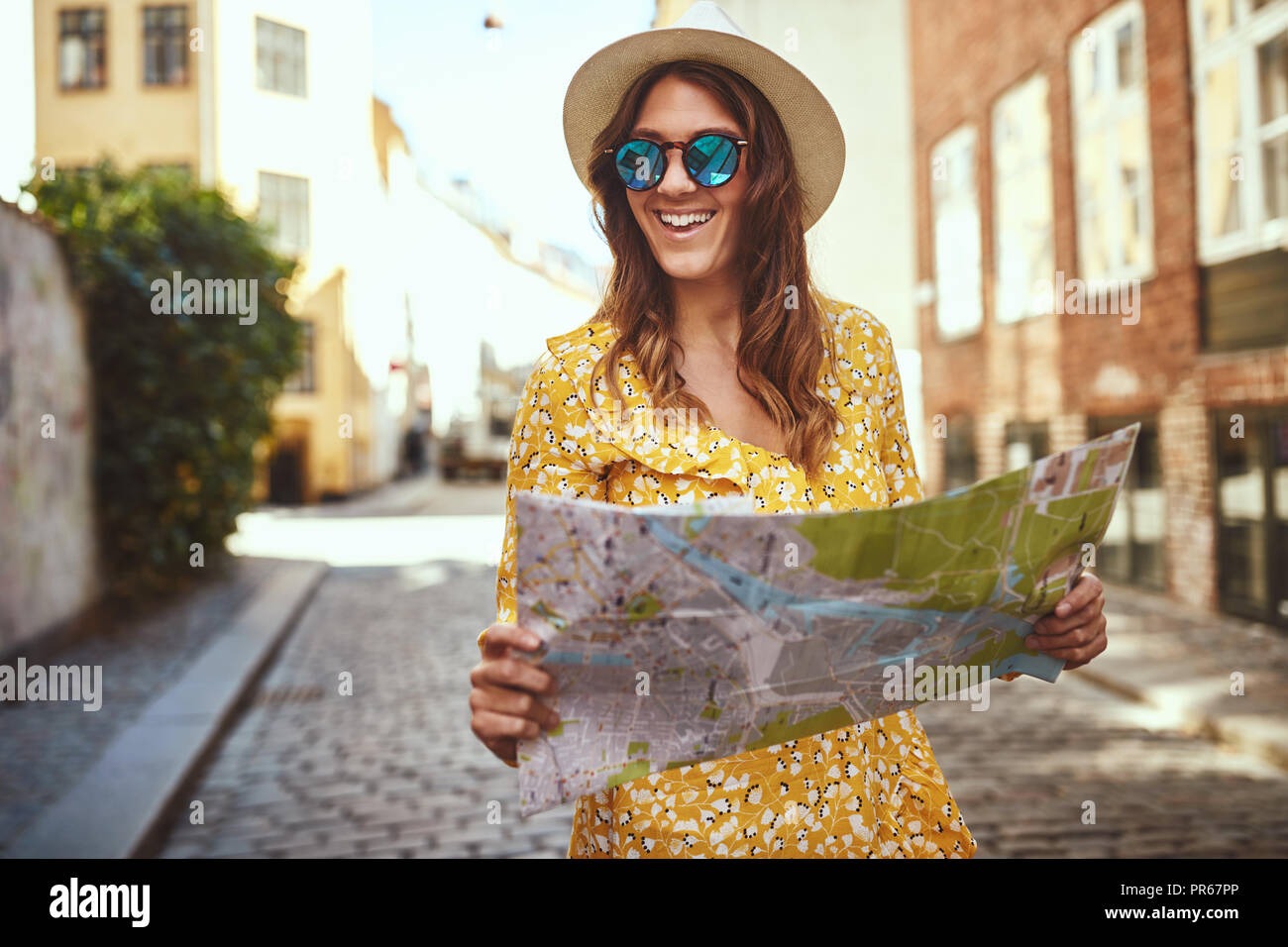 Smiling young brunette woman holding a map while exploring the cobblestone streets of a city wearing a hat and sunglasses Stock Photo