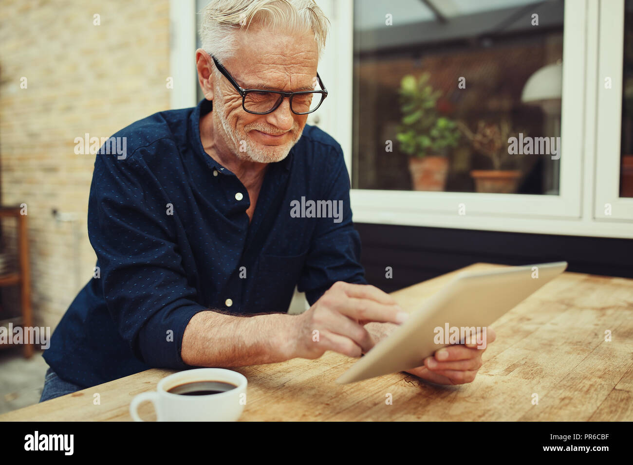 Smiling senior man working nline with a tablet and drinking a cup of coffee while sitting at a table outside on his patio Stock Photo