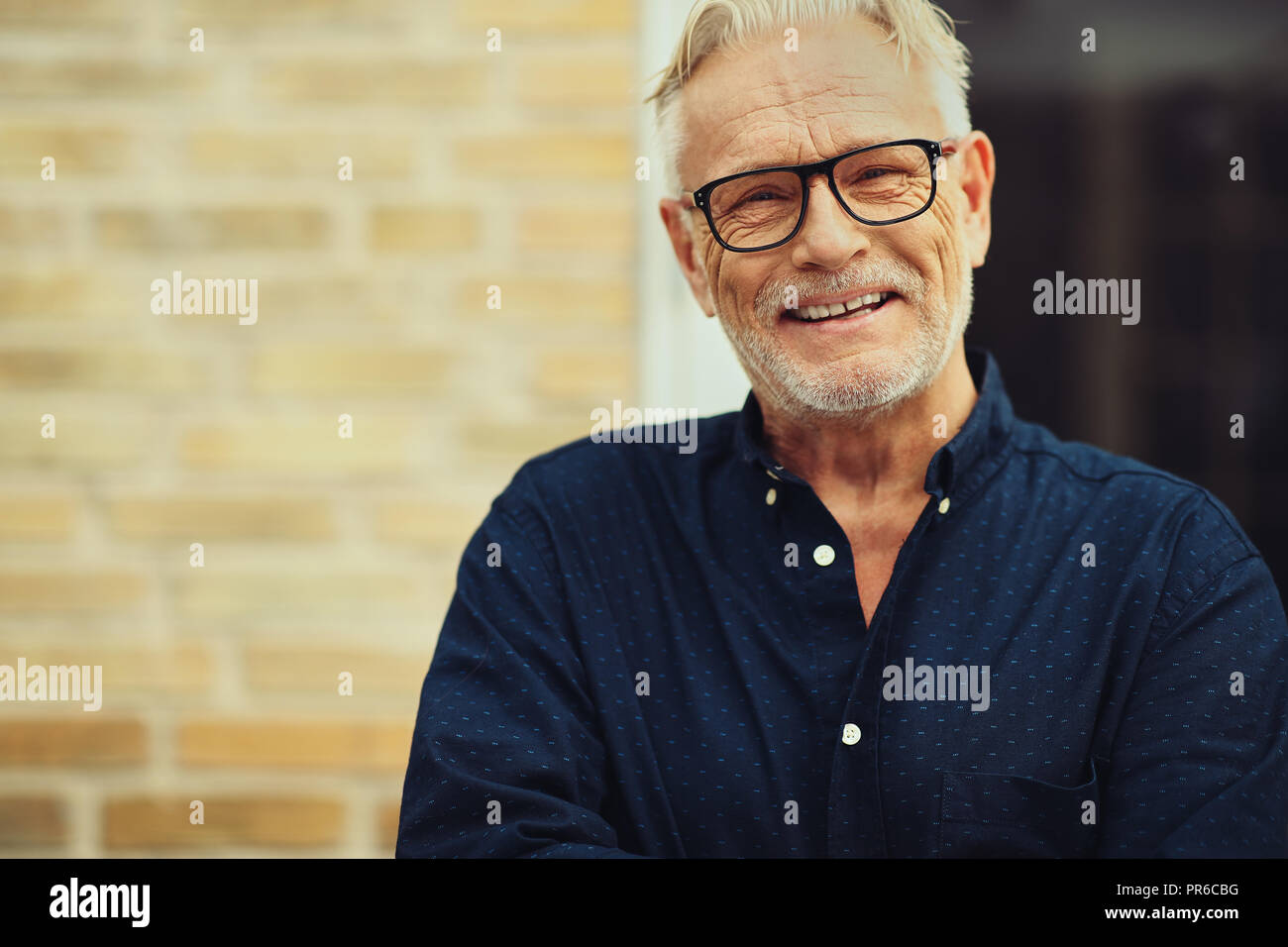 Content senior man with a beard and wearing glasses laughing while standing outside in front of his home Stock Photo