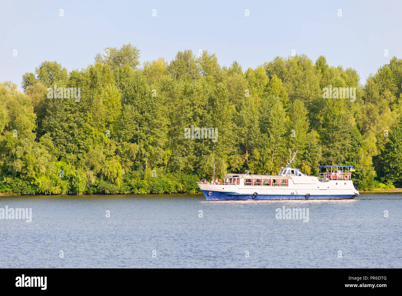A white and blue boat on the Dnieper river, close to the shore with a forest of trees, in Kiev, Ukraine Stock Photo