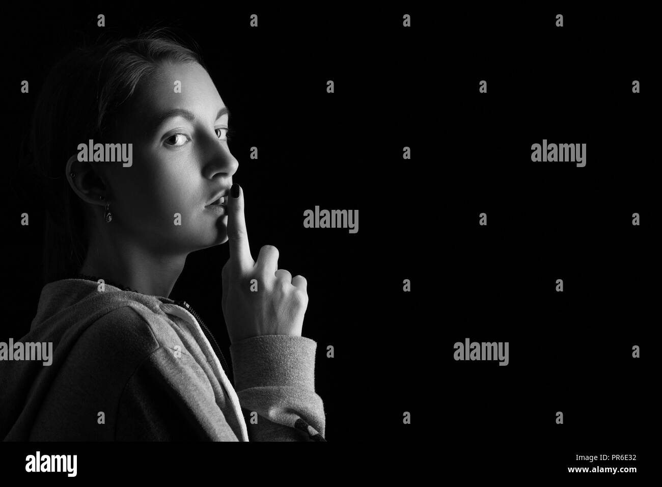 scared girl show silence gesture on black background with copy space, looking at camera, monochrome Stock Photo