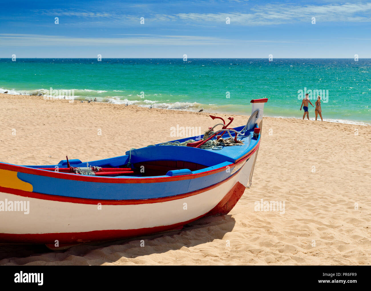 fishing boat painted in traditional colours, on Armacao de Pera beach with a distant couple walking on the beach Stock Photo
