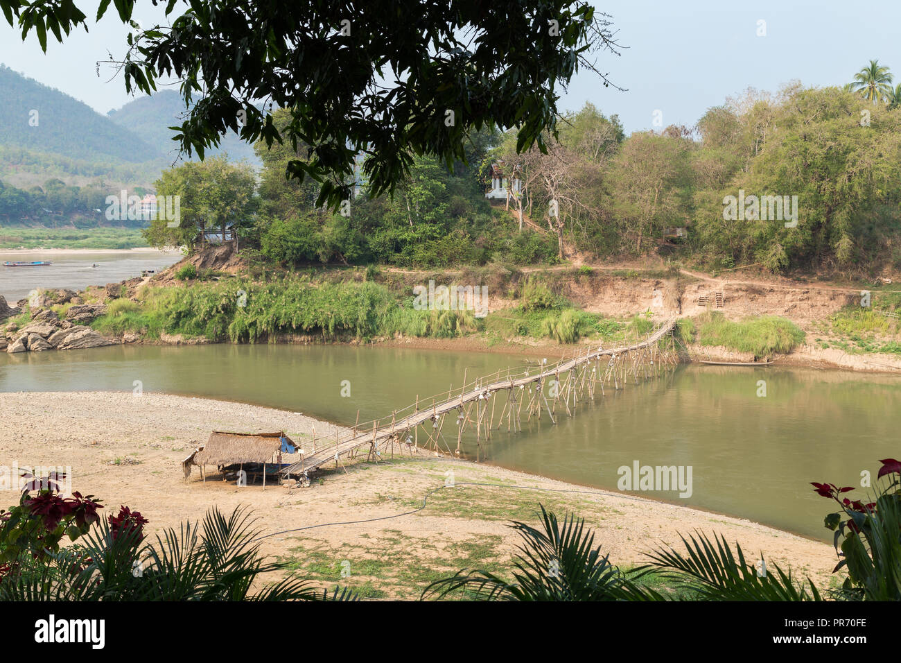 View of a bamboo bridge over Nam Khan River at low tide and lush riverbank in Luang Prabang, Laos, on a sunny day. Stock Photo