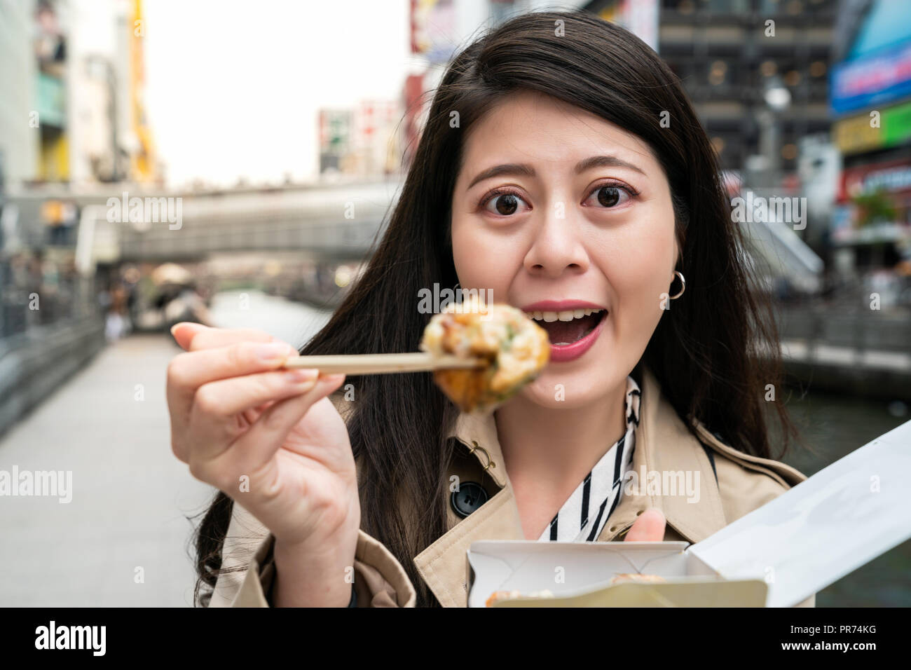 female tourist cheerfully standing next to the river and using chopsticks to eat takoyaki, she is giving a big attractive smile to the camera Stock Photo