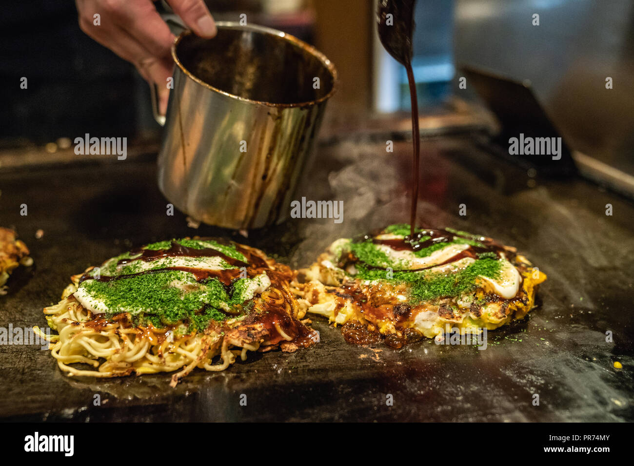 the delicious okonomiyaki is almost done on the iron plate, the cook is adding some japanese traditional sauce Stock Photo